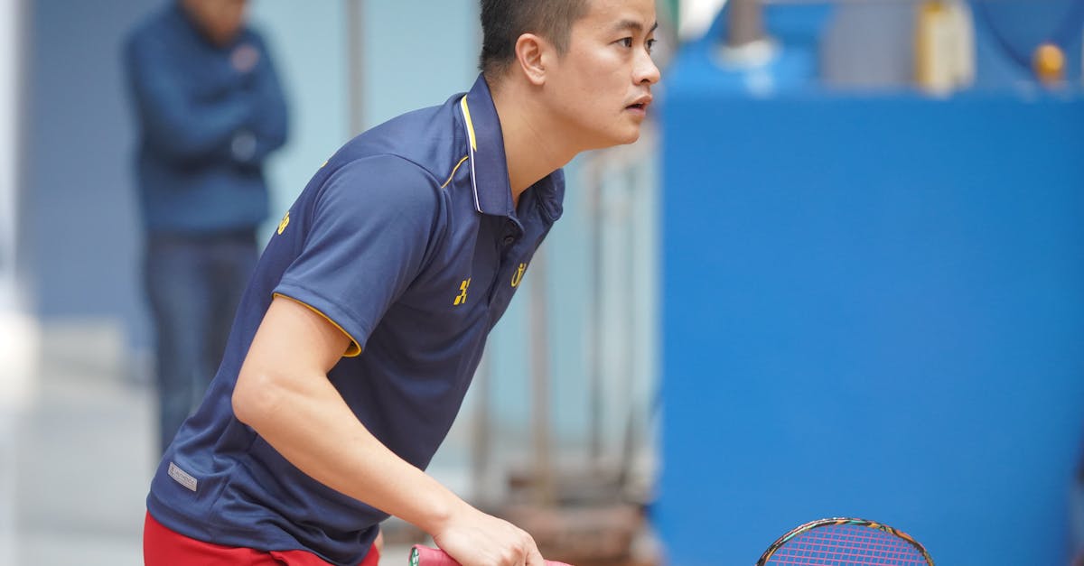 Asian man in blue polo shirt playing badminton indoors, showing concentration and athleticism.