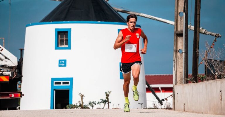 A fit young man in a red jersey jogs energetically near a traditional windmill.