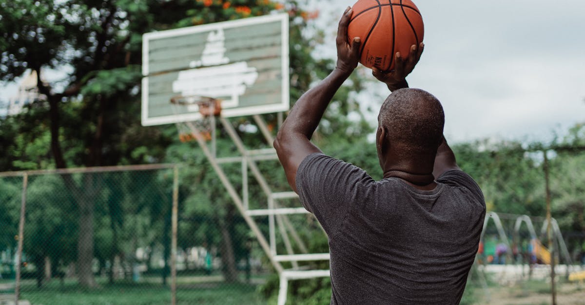 An adult man playing basketball outdoors, aiming for a shot in a lush park setting.