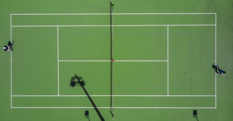 High-angle drone shot of a tennis match on a bright green court with shadows.