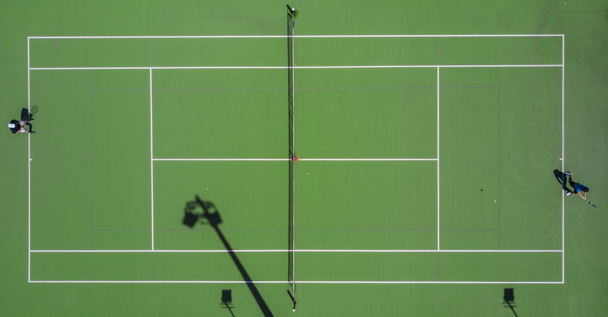 High-angle drone shot of a tennis match on a bright green court with shadows. Ukuran lapangan tenis