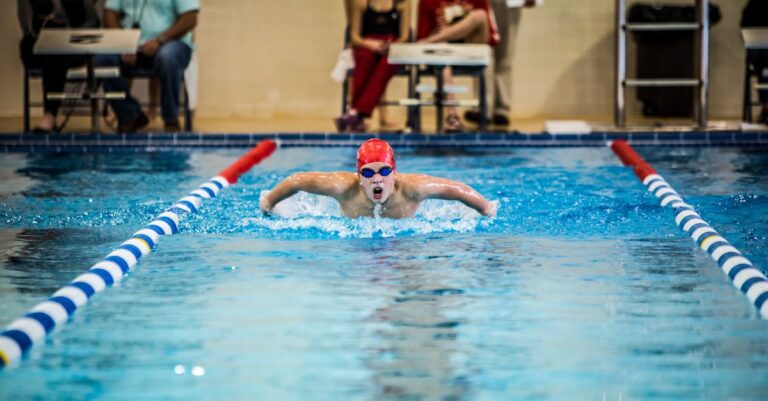 A young swimmer in a red cap competes in a butterfly stroke during an indoor pool race.