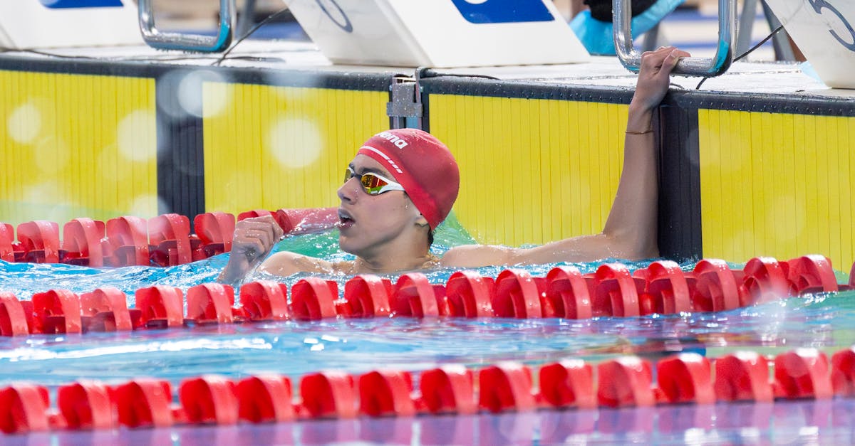 Swimmer in red cap and goggles finishes a race, holding lane divider. Celebrating victory.