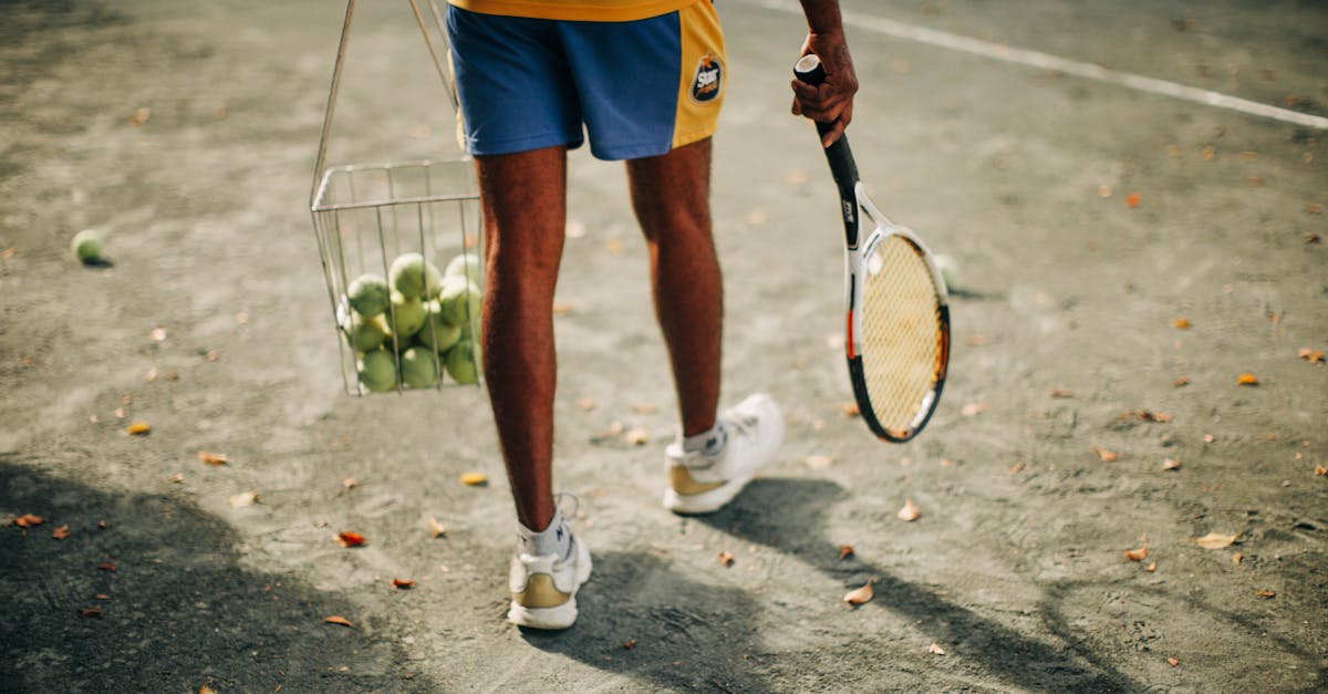 A tennis player stands on a clay court holding a racket and a basket of tennis balls, ready for practice.